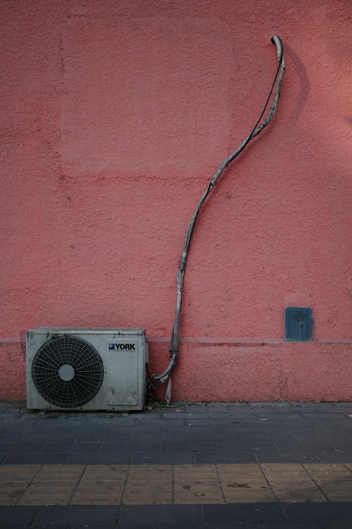 Air conditioning unit against a pink wall with a sidewalk, showing urban setting.