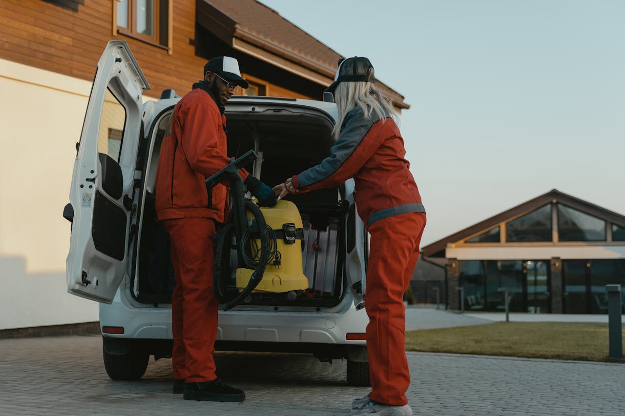 Two cleaners loading a vacuum into a van outside a modern building at dusk.